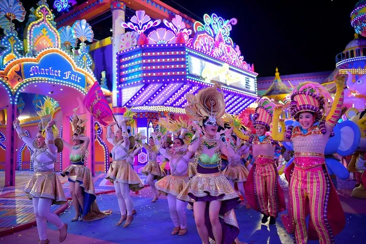 Dancers in sequinned costumes and feathered headdresses parading past neon-lit carnival facades at Carnival Magic, Phuket